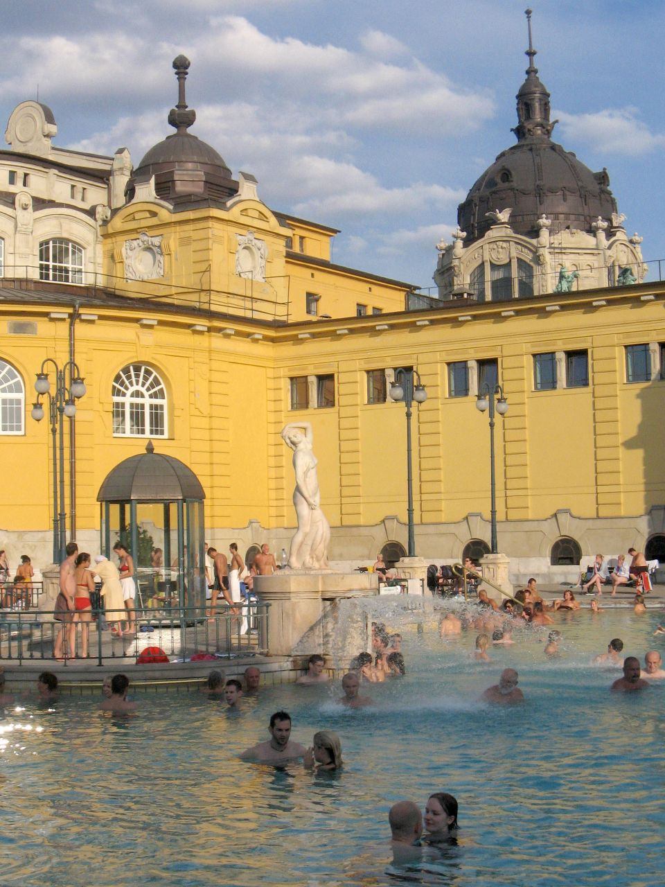 Bathers in the yellow Neo-Baroque outdoor thermal pool courtyard of Szechenyi Baths, Budapest, with the domed main building and a classical fountain statue in the background